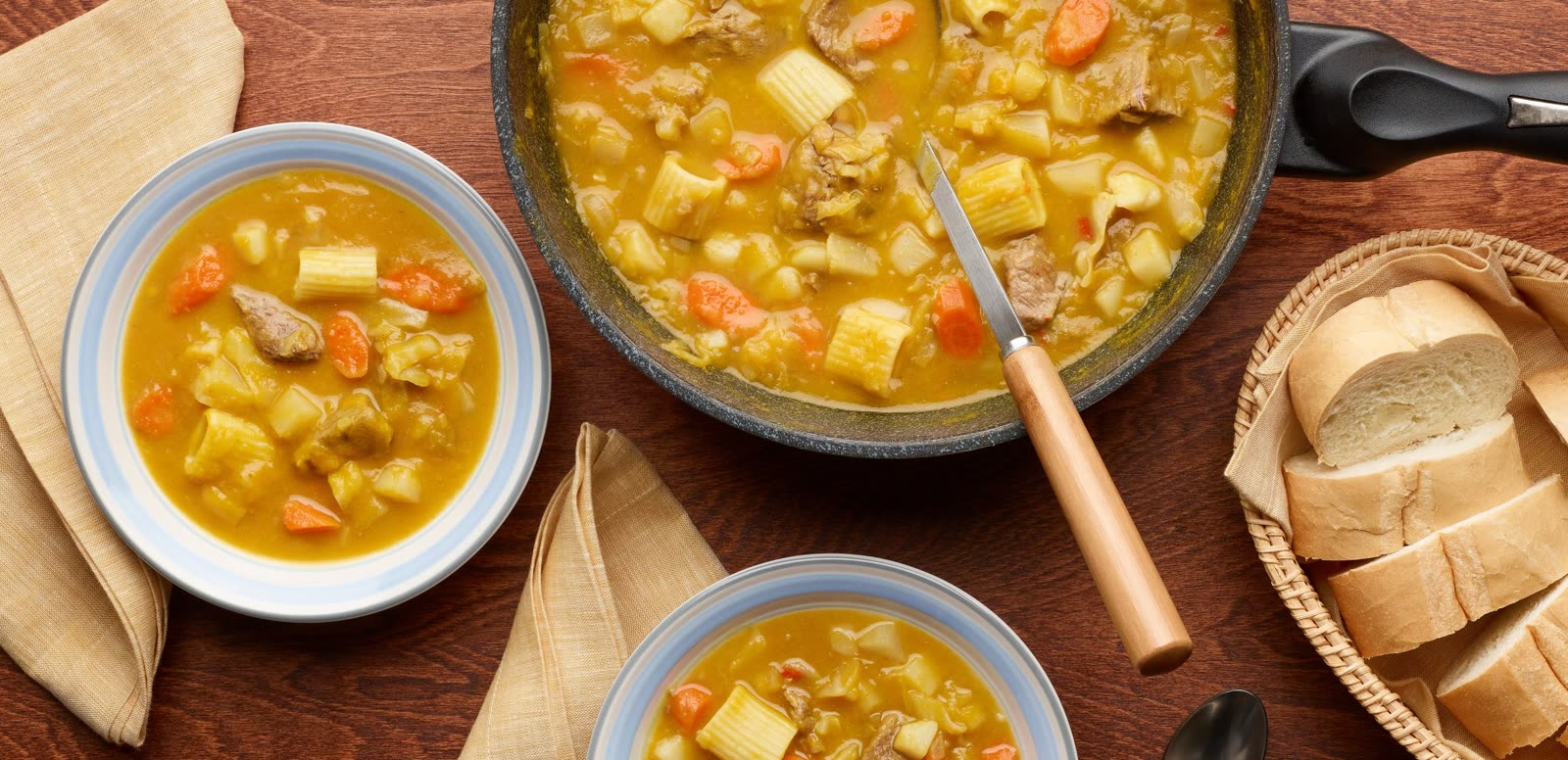 Two bowls and a pot of soup joumou — beef, rigatoni, carrot and potato in a golden squash broth — with sliced bread alongside.